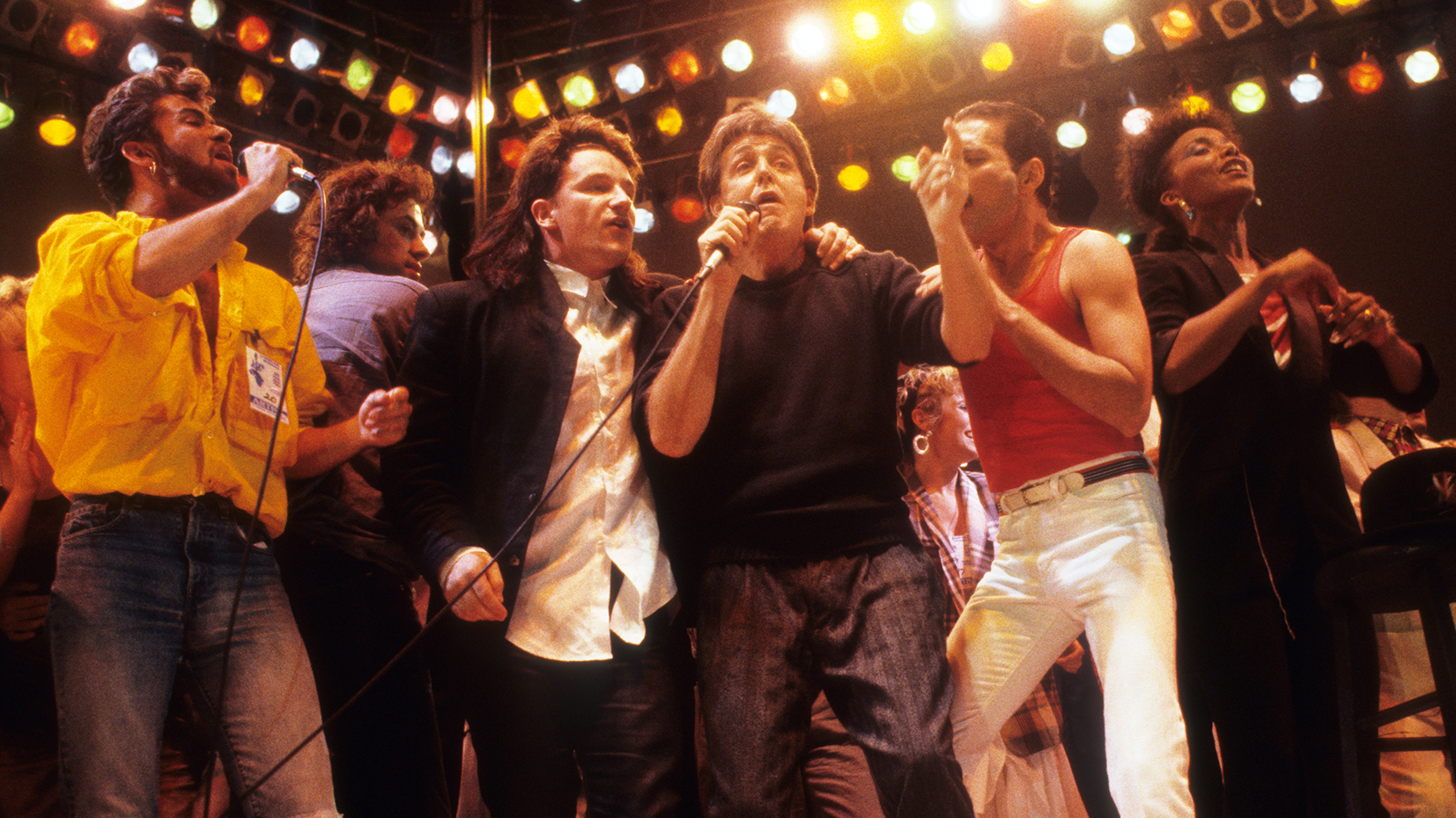 George Michael, Bono, Paul McCartney, Freddie Mercury, and other iconic performers sing together on stage under bright concert lights during the grand finale of Live Aid 1985.