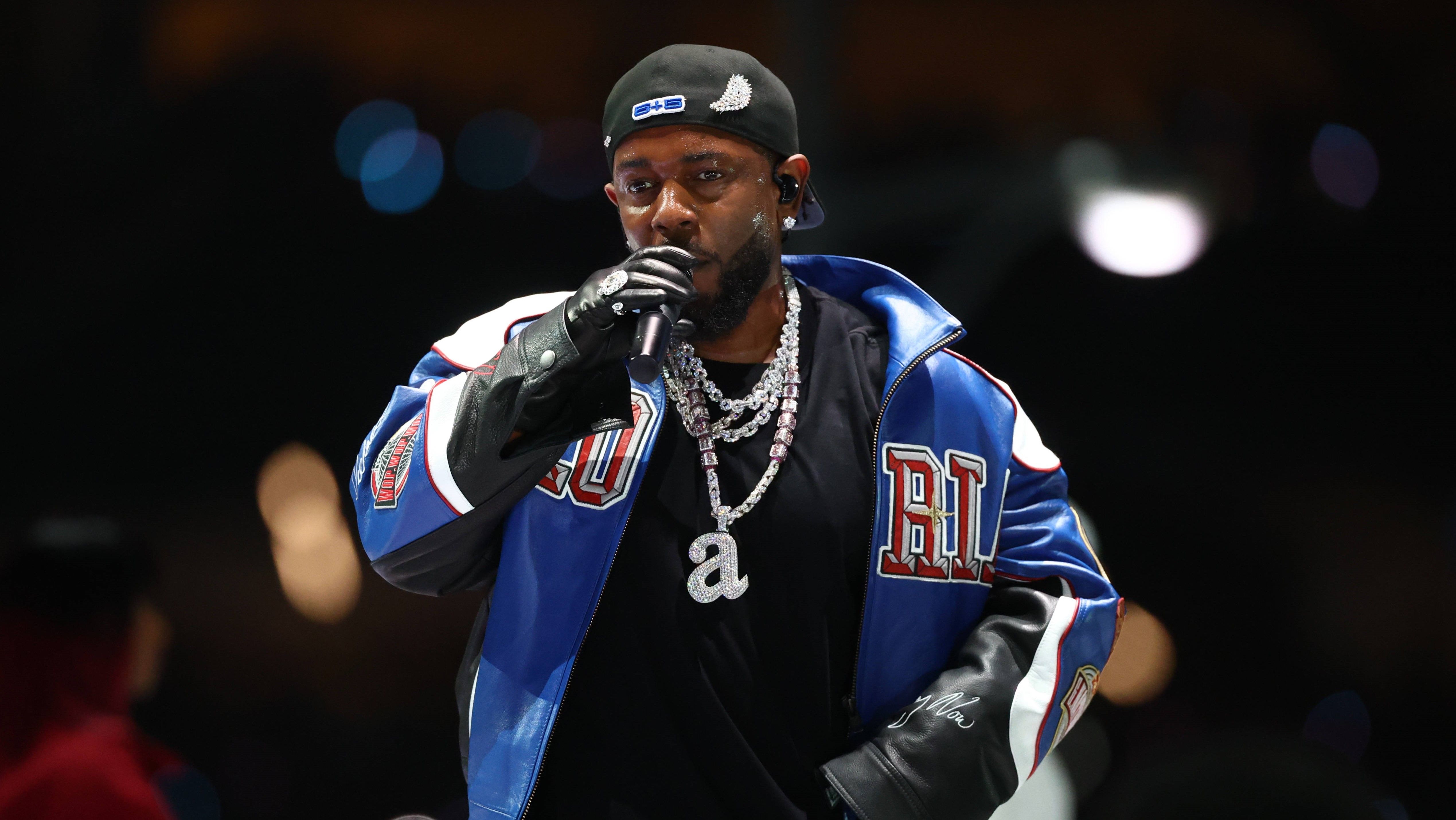 Kendrick Lamar performing on stage wearing a blue and black leather jacket, black gloves, a backwards cap, and layered diamond chains, holding a microphone.