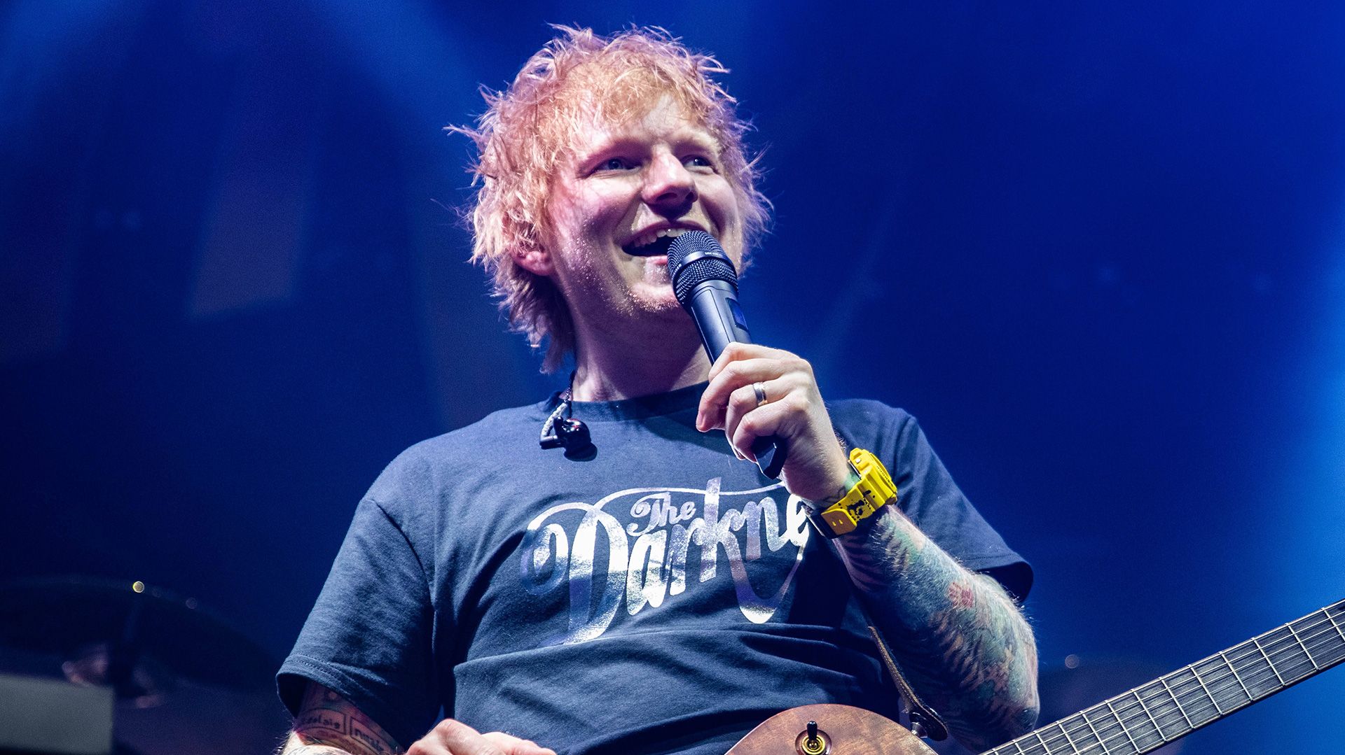 Ed Sheeran wearing a blue T-shirt, speaking into a microphone, with a guitar around his neck.