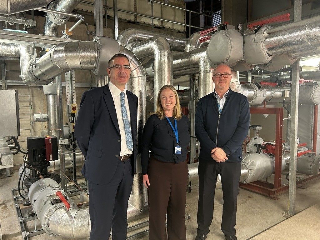 Dr Clive Winters stood next to the head and deputy heat of sustainability in with the new energy heat network behind them made up of large silver pipes